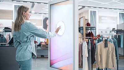Young blonde woman in denim jacket taps on "Press Start" control panel of large advertising and info display for modern shopping experience in fashion store