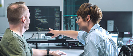 An IT specialist and an IT specialist consult on IT measures in front of several monitors with programming language