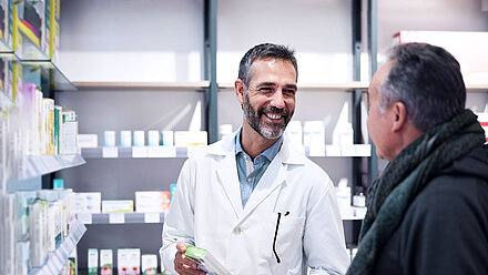 Two men in a pharmacy; Pharmacist with short black hair and beard; wears a doctor's coat and holds a medicine in his hand. He smiles at man standing in front of him, also black short hair, jacket and scarf.