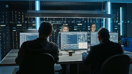 Four people monitor computer screens in a dark server room with blue lighting.