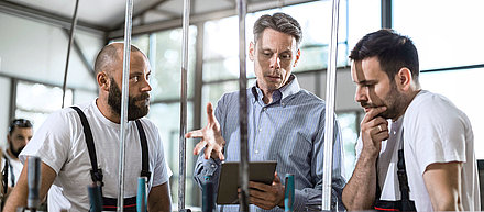 Two young factory employees look at digital evaluations of a machine on a tablet with their older colleague in the production hall and draw valuable conclusions for their production from them