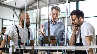 Two young factory employees look at digital evaluations of a machine on a tablet with their older colleague in the production hall and draw valuable conclusions for their production from them