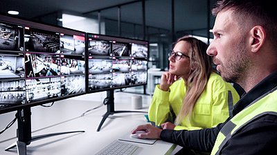 Photo: Two people sitting in front of multiple monitors displaying live camera feeds while monitoring a security and video surveillance system.