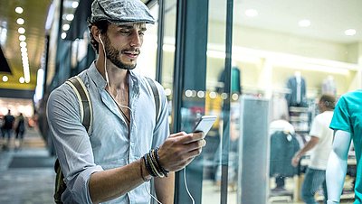 Gentleman in a slouchy cap, shirt, backpack and headphones in his ear looks at his smartphone before entering a store