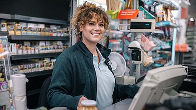 Young cashier with light brown curly hair sits at checkout and smiles in the camera
