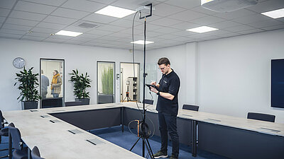 Technician stands in front of a tripod with an access point and looks at a tablet; colleagues can be seen in the background