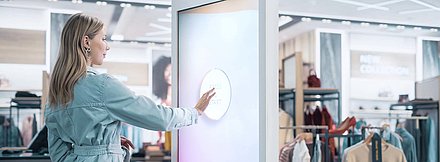 Young blonde woman in denim jacket taps on "Press Start" control panel of large advertising and info display for modern shopping experience in fashion store