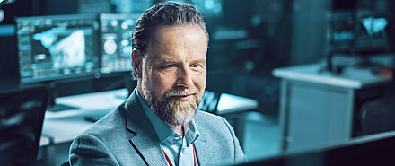 Office worker in suit with PC screens in background smiles reassuringly