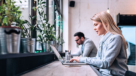 Photo of two people working on their laptops in a modern office or café; on the left, many potted plants are seen on the windowsill; on the right, a young, blonde woman in the foreground and a young, brunette man with glasses in the background are sitting at their laptops facing the window, working on something in a good mood