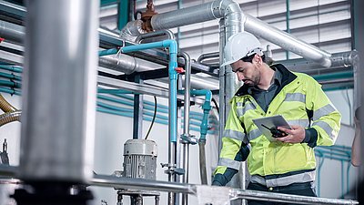 Photo: Technician wearing a safety helmet, high-visibility jacket, and holding a tablet while standing among pipelines and technical plant components in an industrial environment.