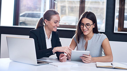 Civil servant in pigtail and blazer sits at desk with colleague and shows her how to fill out a digital form on a tablet