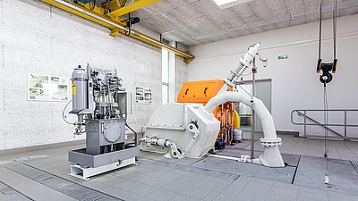 Industrial turbine system with hydraulic unit in a bright technical room, ceiling-mounted crane hook visible.