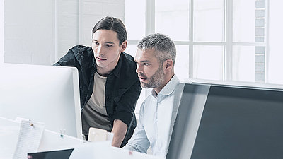 Young IT employee with pigtail advises middle-aged colleague with gray hair and beard on PC security settings