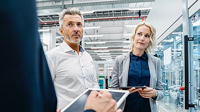 Photo: Two people with tablets standing in a modern industrial or production environment with glass-enclosed machinery areas and technical equipment.