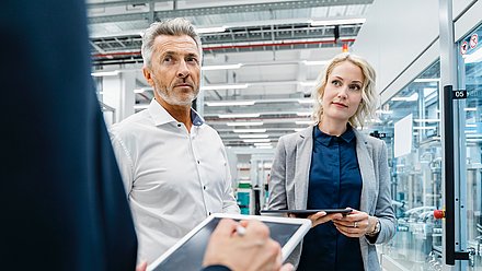 Photo: Two people with tablets standing in a modern industrial or production environment with glass-enclosed machinery areas and technical equipment.