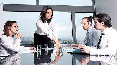 Photo of a group of four office workers discussing a document in front of a window in a conference room in business attire: A young brunette woman confidently points to the document on the table