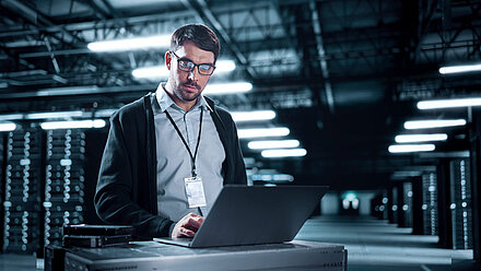 Man with glasses works on a laptop in a lit server room with data racks in the background
