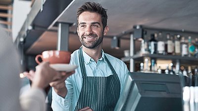 Friendly barista at cash register with beard and apron smilingly hands coffee cup to customer
