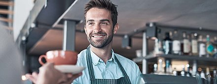 Friendly barista at cash register with beard and apron smilingly hands coffee cup to customer