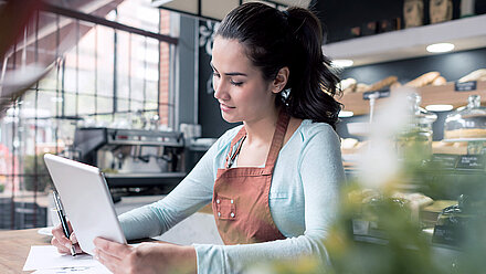 Female employee of a bakery is sitting at a table, holding a tablet and a pen, with various documents in front of her