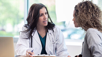 Young doctor with long brown hair and a doctor's coat explains something to a patient with light brown curls at the consultation table in the doctor's office