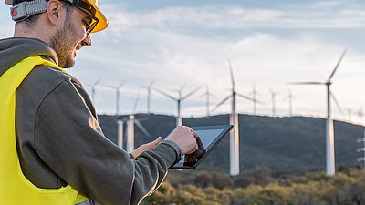 Photo: Person wearing a safety helmet and high-visibility vest operating a tablet in front of several wind turbines in a hilly landscape.
