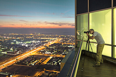 Photo of a man taking a picture of the illuminated city at night with his camera tripod from the balcony / roof of his penthouse apartment above a big city