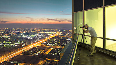 Photo of a man taking a picture of the illuminated city at night with his camera tripod from the balcony / roof of his penthouse apartment above a big city