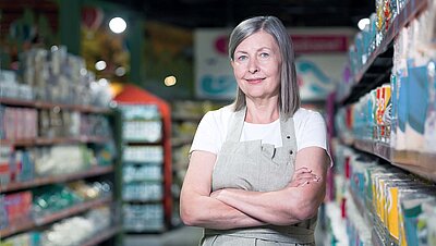 Middle-aged store manager with gray long hair and work apron stands between two supermarket shelves smiling contentedly