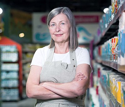 Middle-aged store manager with gray long hair and work apron stands between two supermarket shelves smiling contentedly