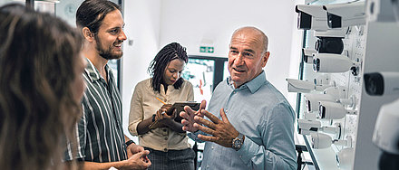 Middle-aged gentleman stands in front of wall with white camera heads and enthusiastically tells younger colleagues and visitors about his products