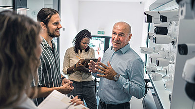 Middle-aged gentleman stands in front of wall with white camera heads and enthusiastically tells younger colleagues and visitors about his products