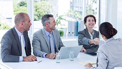 Contract signing scene in white office building: rear view of brunette woman with ponytail and jacket shaking the hand of another short-haired woman in business attire with a friendly smile; next to her, two smiling colleagues in suits with laptops sit at the same table and watch the two women with satisfaction