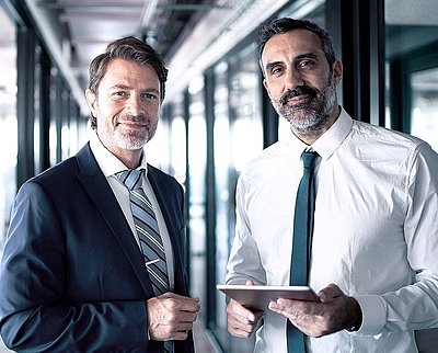 Two businessmen in suits and holding tablets stand in office buildings and look confidently into the camera