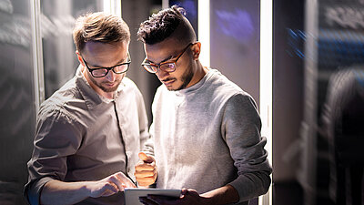 Two men stand in a server room and analyze data together on a tablet.