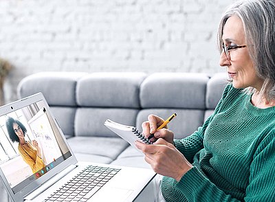 Older lady with gray hair, glasses, and green sweater sits at coffee table with pen and paper in front of laptop and notes important information from a webinar