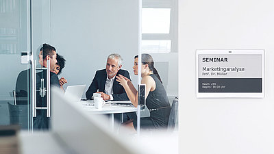 A group of office workers converses animatedly in a conference room labeled with an electronic door sign