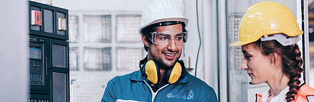 Black-haired worker in utility plant with white hard hat, safety goggles and yellow noise protection headphones in blue overalls and work gloves holding laptop and standing to the right of a control box (left); he explains something to his brunette colleague to his right, smiling; his colleague is also wearing a yellow hard hat and safety vest and listening attentively