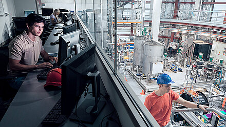 Two-part image from a municipal utility company: on the left, two employees can be seen in the control room at their computers, concentrating on checking values on their screens, while on the right, large tanks, pipes and pumps can be seen behind a pane of glass in a large, bright plant room, as well as a young employee in a construction helmet climbing a staircase