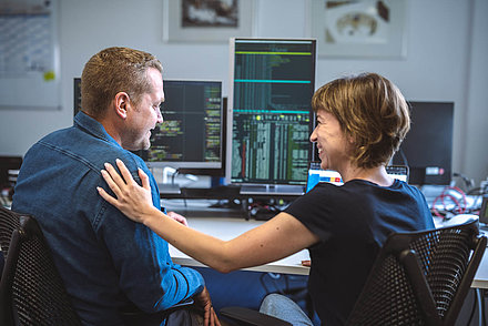 Work situation with view from behind over the shoulders of two employees: Young woman in T-shirt with short, caramel-colored hair pats her blond colleague in shirt appreciatively on the shoulder; in the background, several PC screens with programming language on them can be seen on a desk