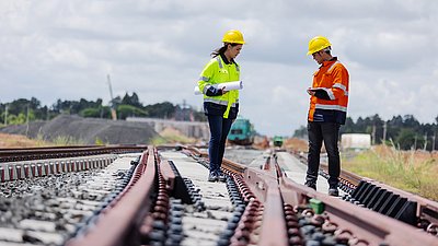 Photo: Two people wearing safety helmets and high-visibility clothing stand on railway tracks and review documents or a tablet.