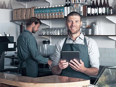 Happy barista in apron stands behind coffee and wine bar with colleagues, holding tablet in hand