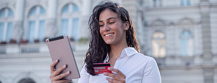 Young woman with curly hair and blouse stands in front of administration building and, with her bank card in hand, contentedly completes a digitalized administration process on her tablet