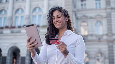 Young woman with curly hair and blouse stands in front of administration building and, with her bank card in hand, contentedly completes a digitalized administration process on her tablet