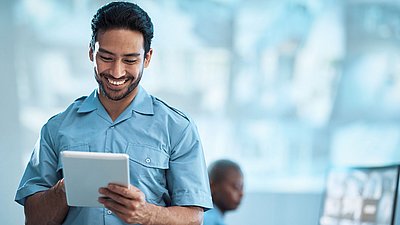 Photo: Security employee in blue workwear using a tablet for the mobile control and monitoring of integrated security and partner solutions in a modern critical infrastructure environment.