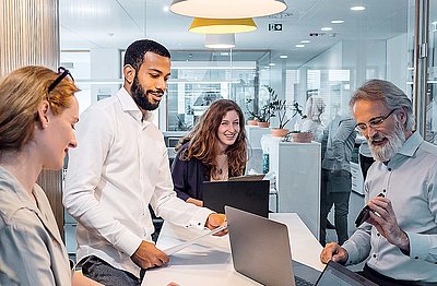 Friendly office worker with a full gray beard and elegant shirt shows his younger colleagues in the office something inspiring on the smartphone and makes the group laugh