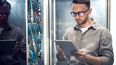 Young IT employee with glasses checks security values on tablet in server room