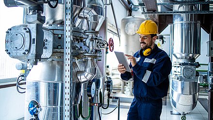 Photo: Technician wearing a safety helmet and holding a tablet while standing among industrial pipelines, valves, and technical plant components.
