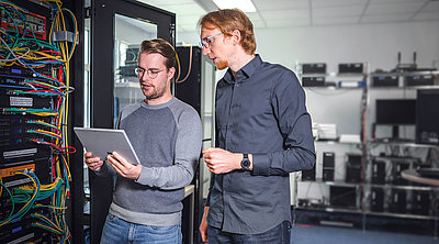 Two men stand in front of a server cabinet and discuss