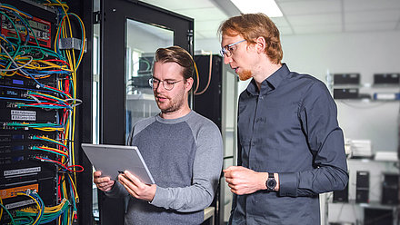 Two men stand in front of a server cabinet and discuss